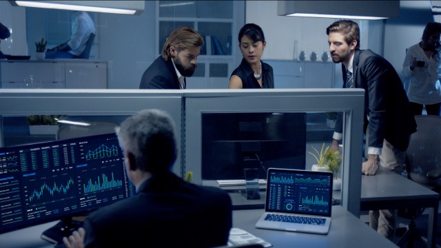 Diverse team of professionals working late at night discussing stock data on computer.